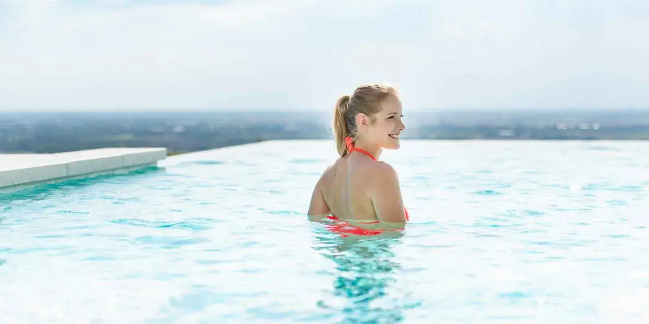 Frau genießt das Schwimmen in einem Infinity Pool, der über den Horizont zu schweben scheint. Was ist ein Infinity Pool? Ein Pool, dessen Wasser über den Rand zu fließen scheint und eine Illusion von grenzenlosem Wasser erzeugt.