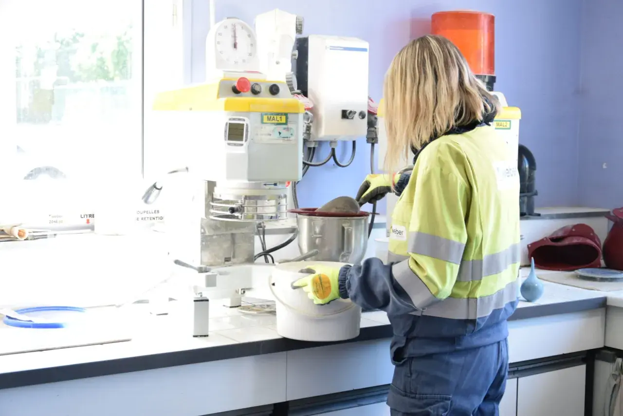Trabajadora en laboratorio, usando guantes, manipula un recipiente con material. Se investiga qué es la silicona y sus usos en la industria.