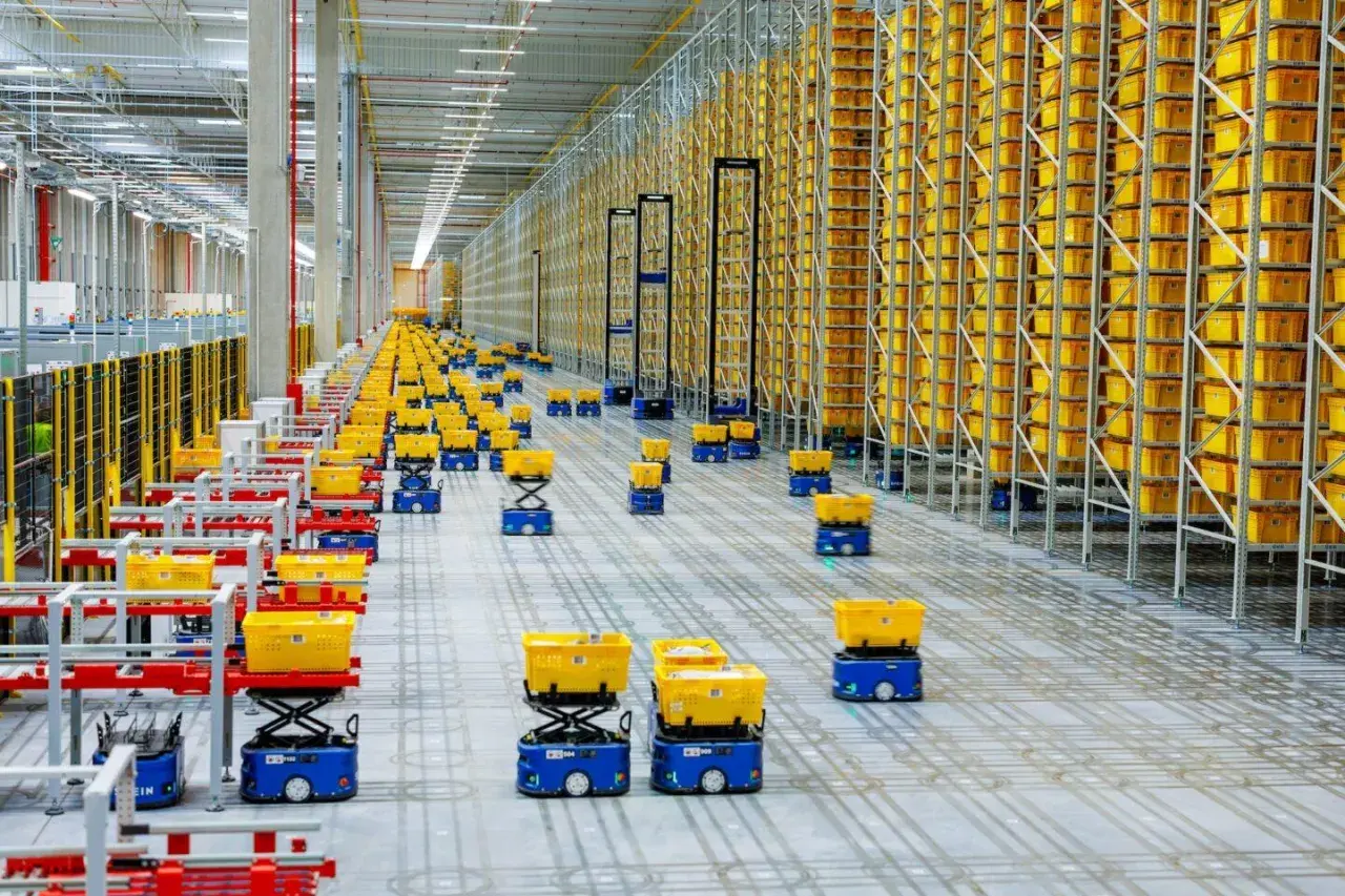 Nowoczesny hub logistyczny z autonomicznymi robotami przewożącymi żółte kosze pośród wysokich regałów wypełnionych towarami.
