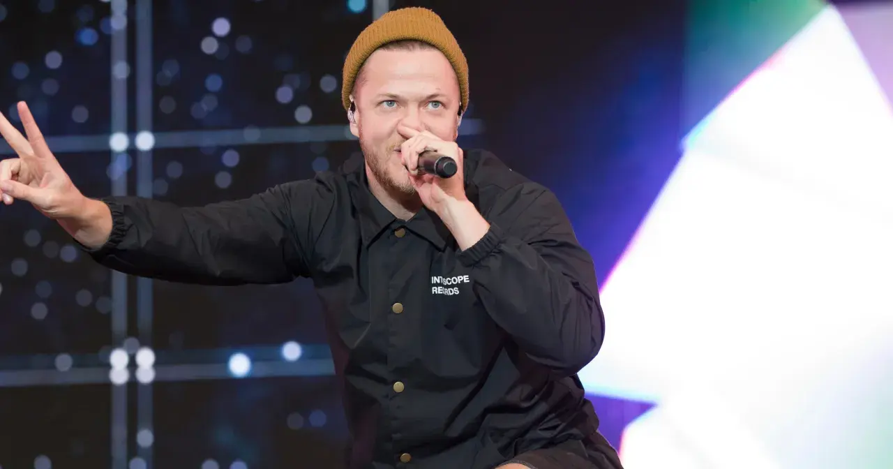 Imagine Dragons wokalista, Dan Reynolds, w beżowej czapce i czarnej kurtce, śpiewa do mikrofonu, pokazując znak pokoju.
