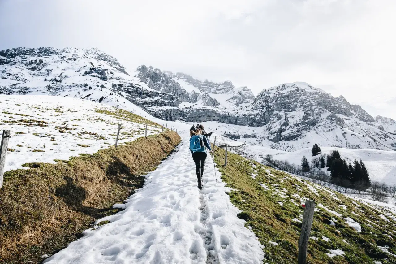 Une randonnée ce week-end dans un paysage montagneux enneigé. Des randonneurs suivent un sentier bordé de clôtures en bois, avec des sommets imposants en arrière-plan.
