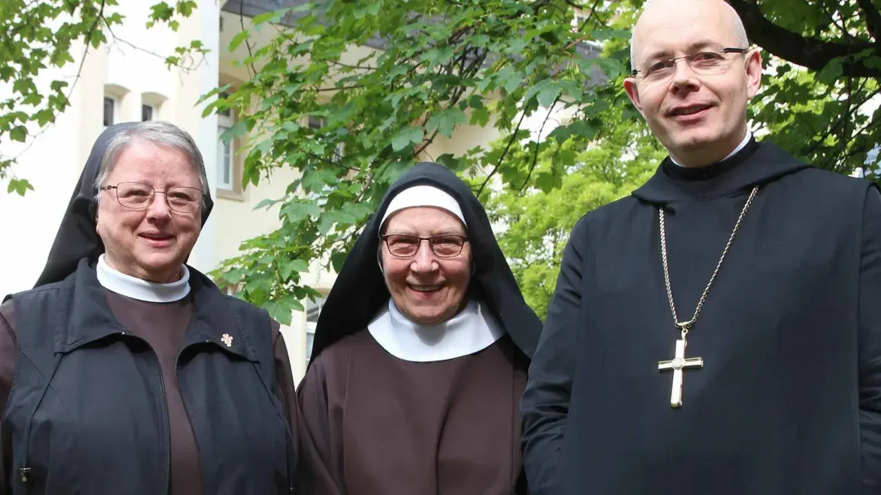 Drei Geistliche, zwei Nonnen und ein Priester, stehen lächelnd vor einem Gebäude. Sie repräsentieren katholische Orden.