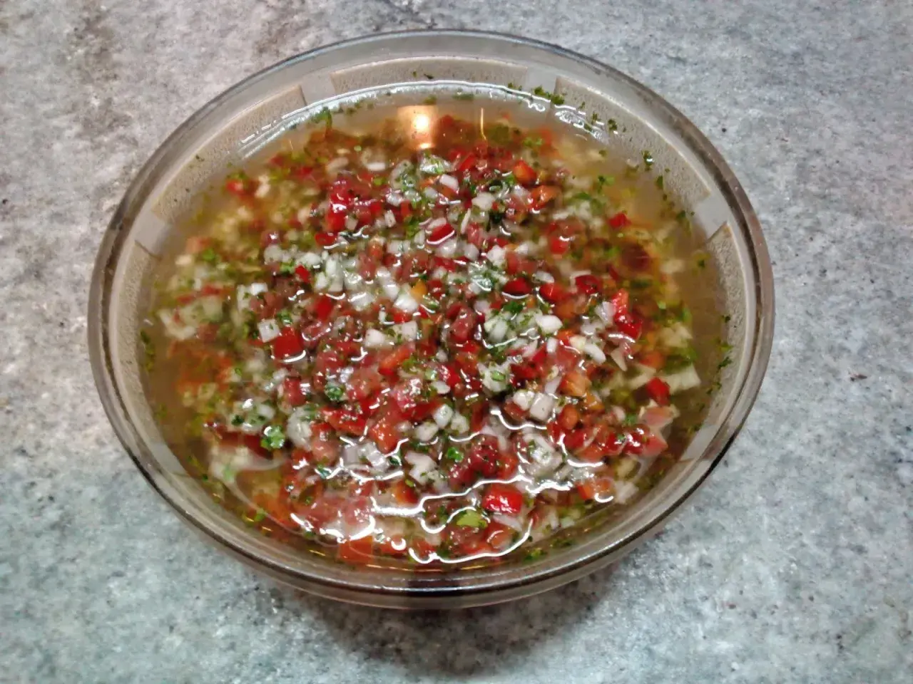 Un tazón de salsa criolla, con trozos picados de tomate, cebolla y pimiento rojo, flotando en un líquido claro.