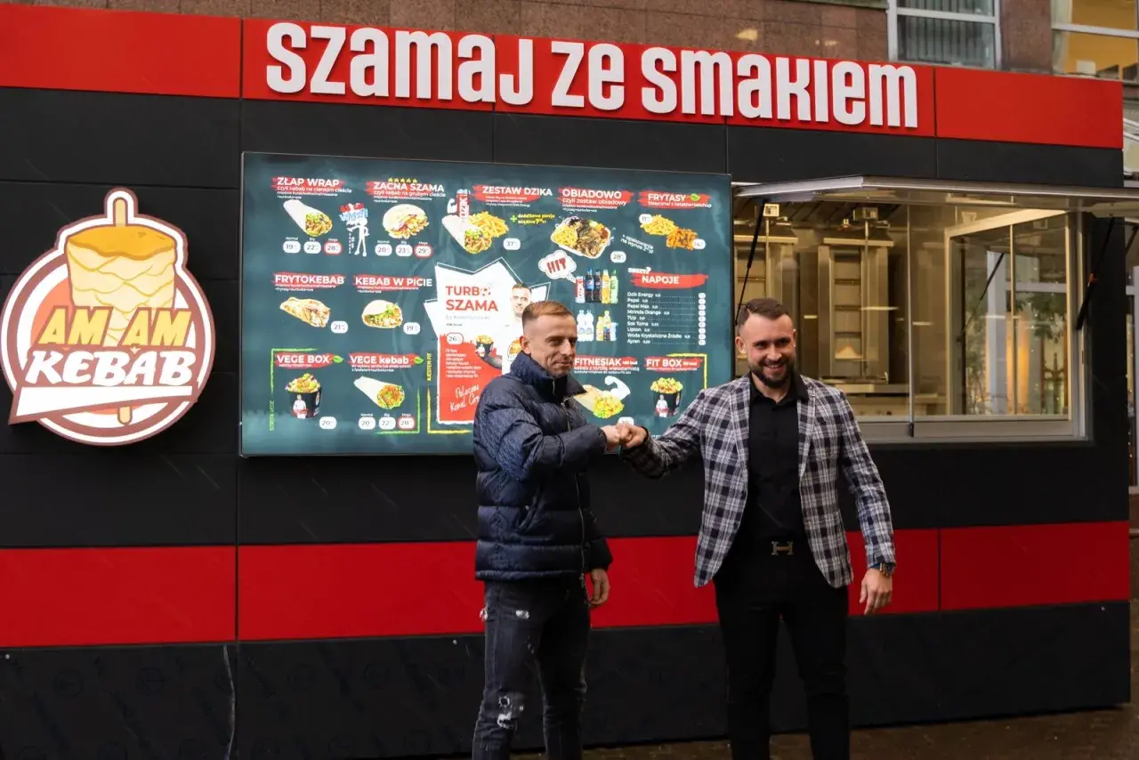 Dwóch mężczyzn wita się przy budce z kebabem, na której widnieje menu.
