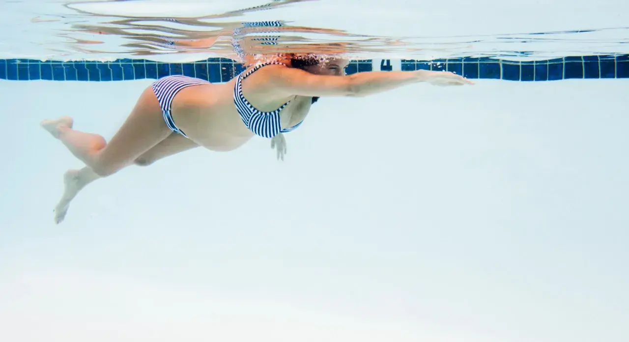En gravid kvinna i randig bikini simmar framåt i en pool. Vattnet är klart och blått.