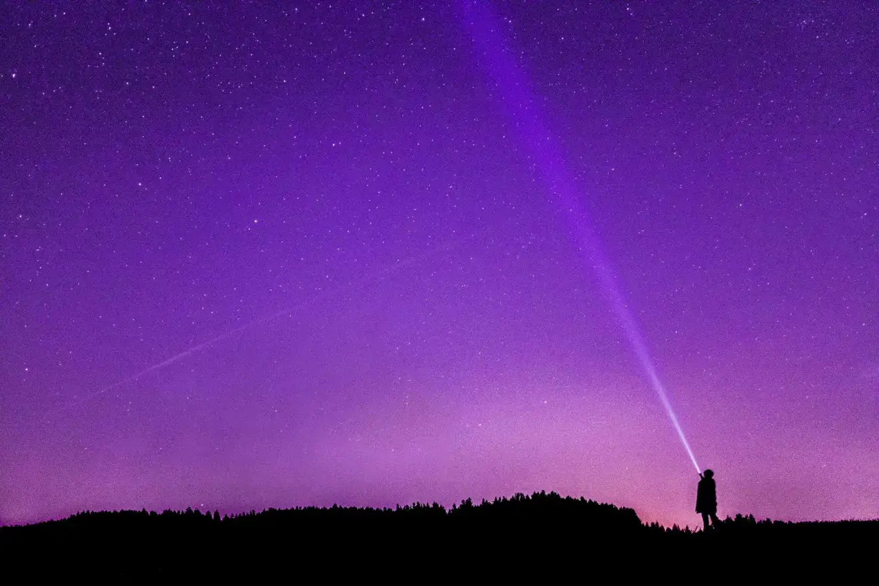 Sylwetka człowieka oświetla latarką nocne niebo, na którym widać gwiazdy i **gwiazdozbiór Tarczy**.