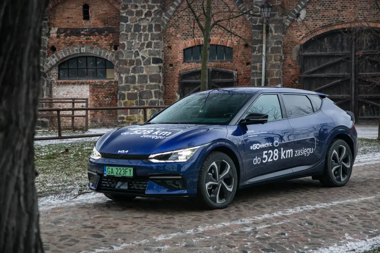 Niebieska Kia EV6 z napisem "#GOelectric D do 528 km zasięgu" stoi przed zabytkową ceglaną budowlą.