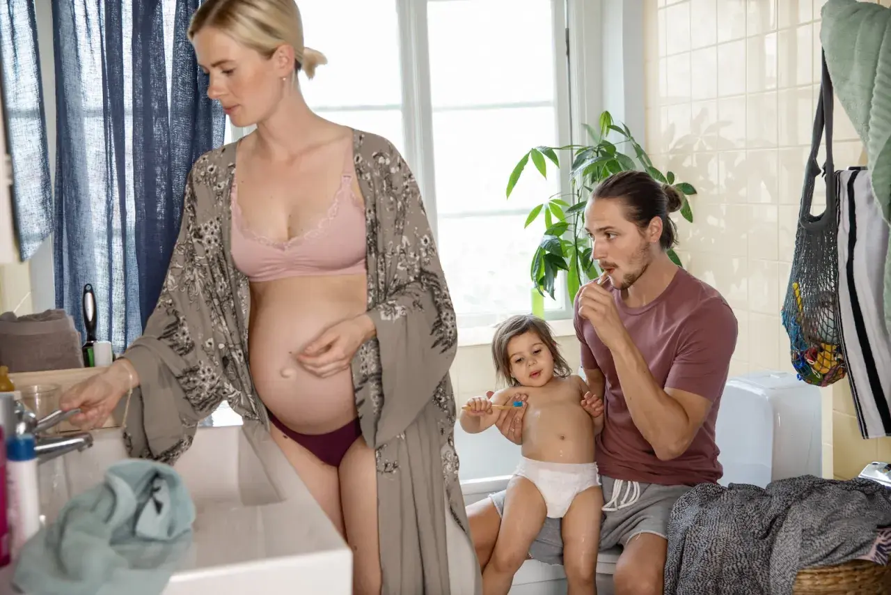 Gravid kvinna vid handfatet, en man borstar sitt barns tänder. En familj förbereder sig för dagen.