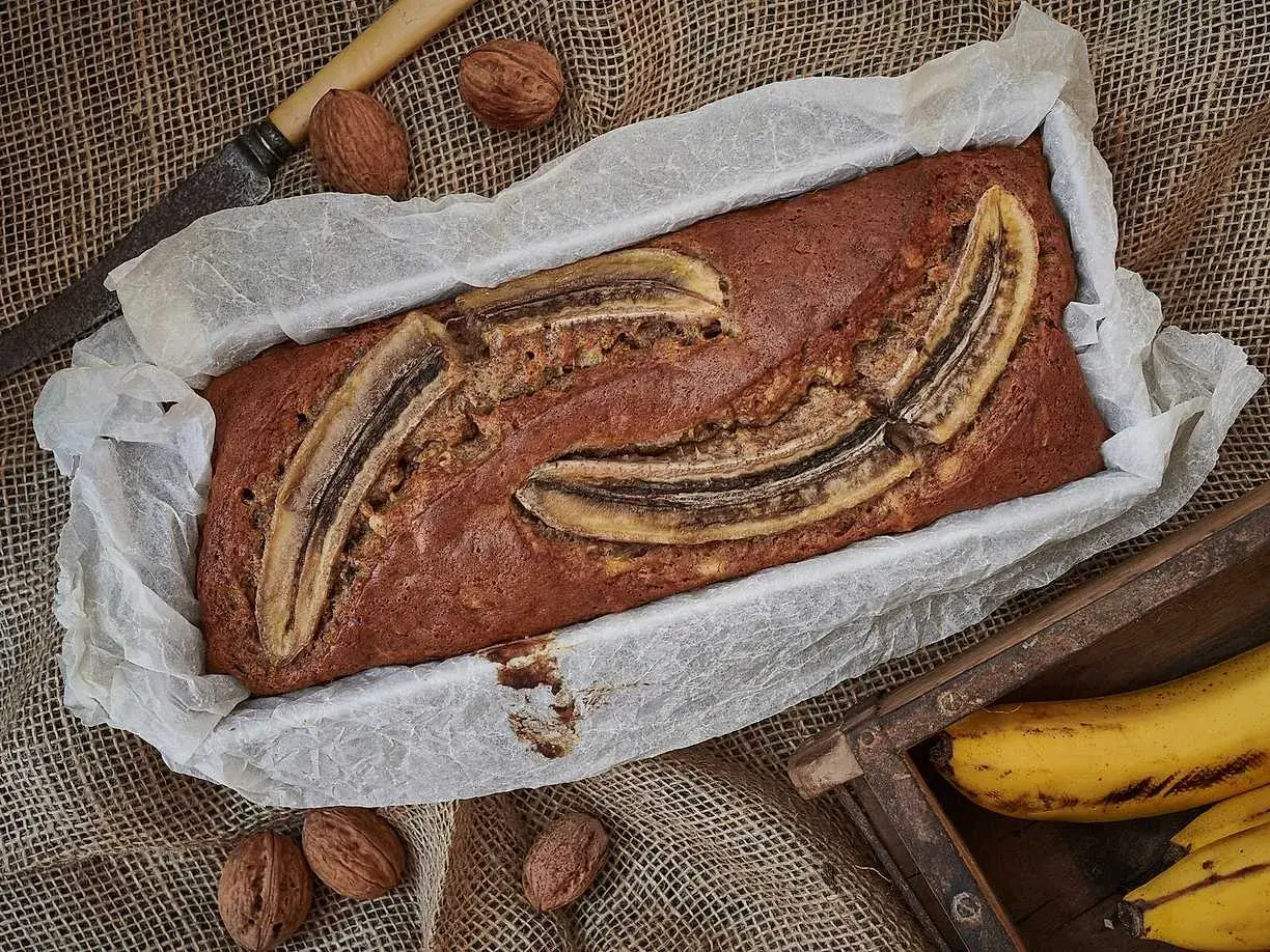 Nybakad banankaka i form, toppad med bananskivor. Valnötter och en gammal kniv ligger bredvid. Perfekt att baka med banan!