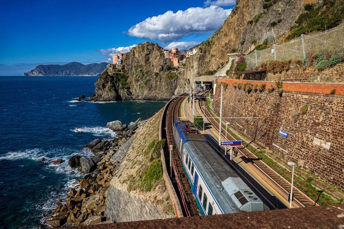 Ein Zug fährt entlang der Küste Italiens am Meer entlang. Bunte Häuser kleben an steilen Felsen.
