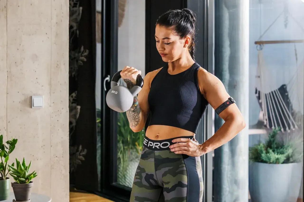Kobieta ćwiczy z kettlebell, co przyspiesza metabolizm. Na ramieniu ma opaskę monitorującą trening.