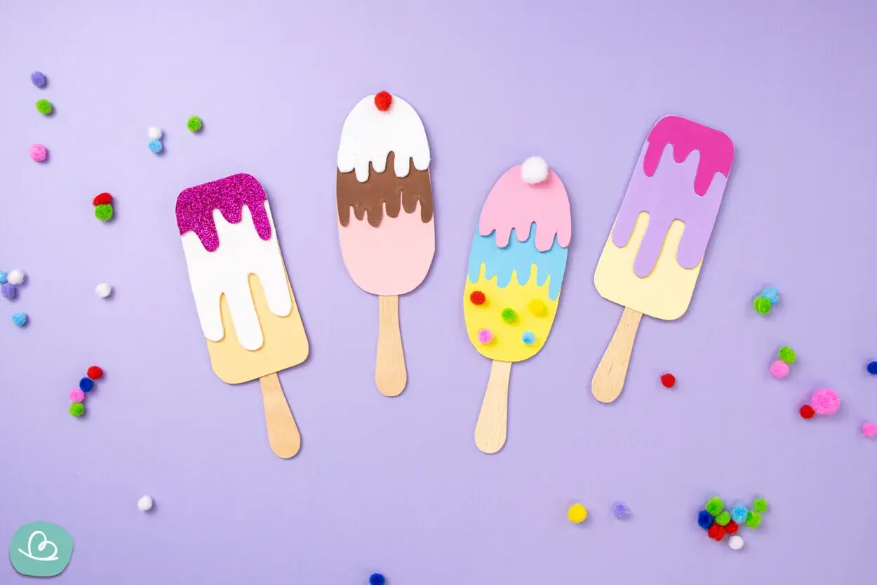 Bunte Eis-Kreationen zum Eis basteln, verziert mit Glitzer und Pompons, liegen auf lila Untergrund.
