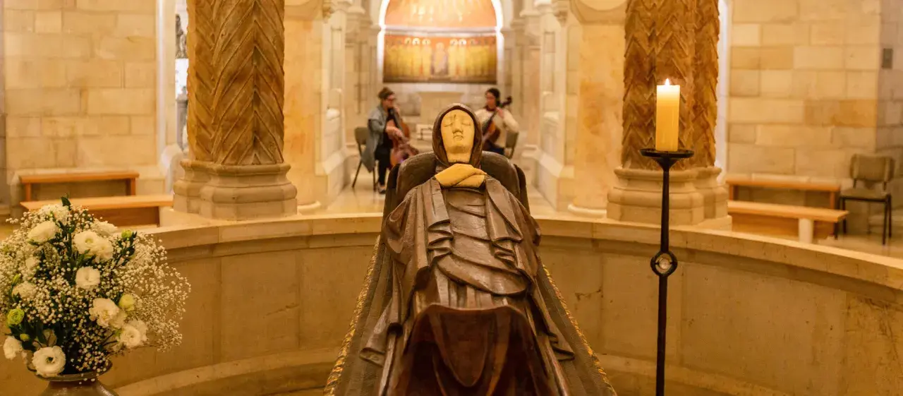 Holzschnitzerei der Mutter Gottes in einer Kapelle, mit Kerzenlicht und Blumen geschmückt. Im Hintergrund spielen zwei Musikerinnen Cello.