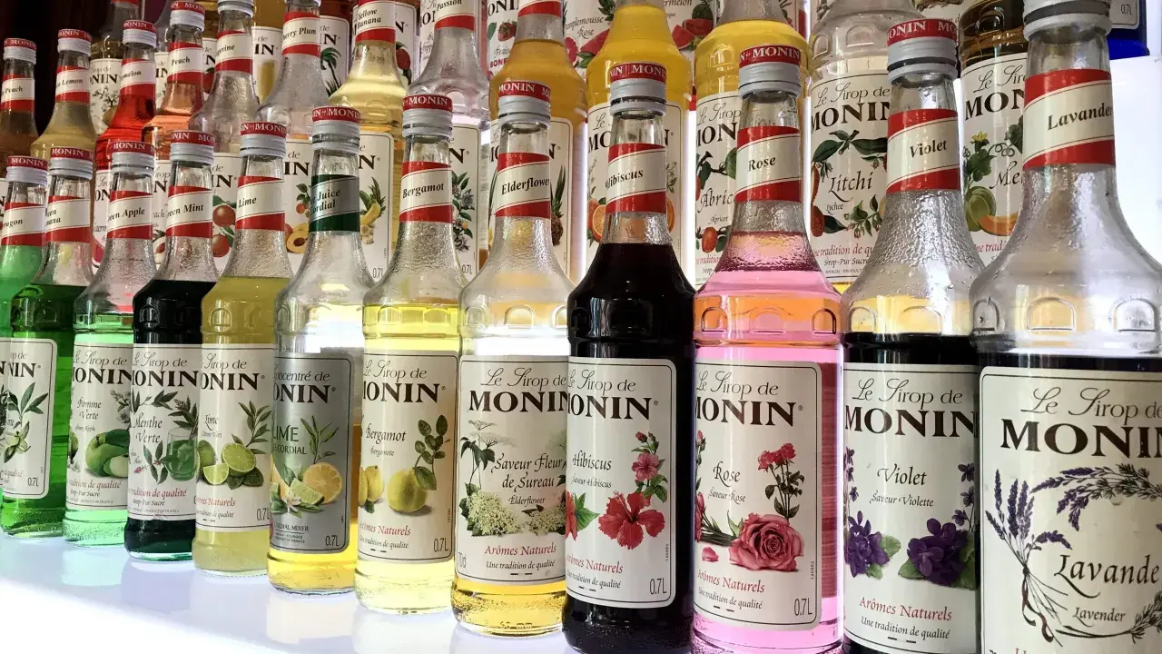 Rząd butelek syropów do kawy Monin o różnych smakach: od owocowych po kwiatowe, np. róża, lawenda, hibiskus.