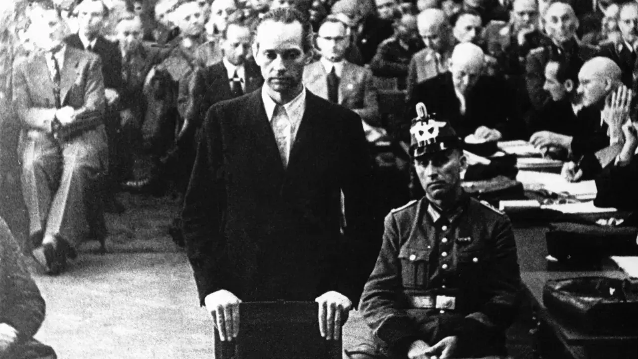 Mann im Anzug vor Gericht, umgeben von Zuschauern. Die Atmosphäre erinnert an die harten Urteile, die Roland Freisler fällte.