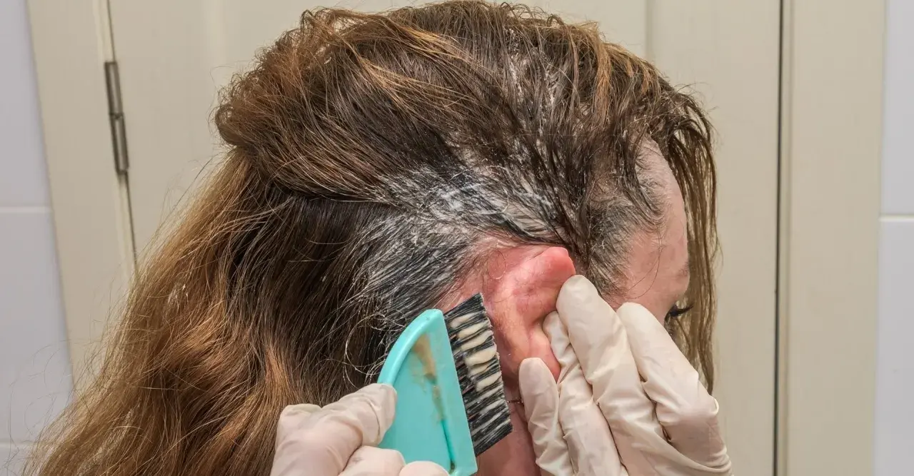 Manos enguantadas usan un cepillo para aplicar tinte en el cabello. Si te manchas la piel, aquí te explicamos como quitar el tinte de la piel.
