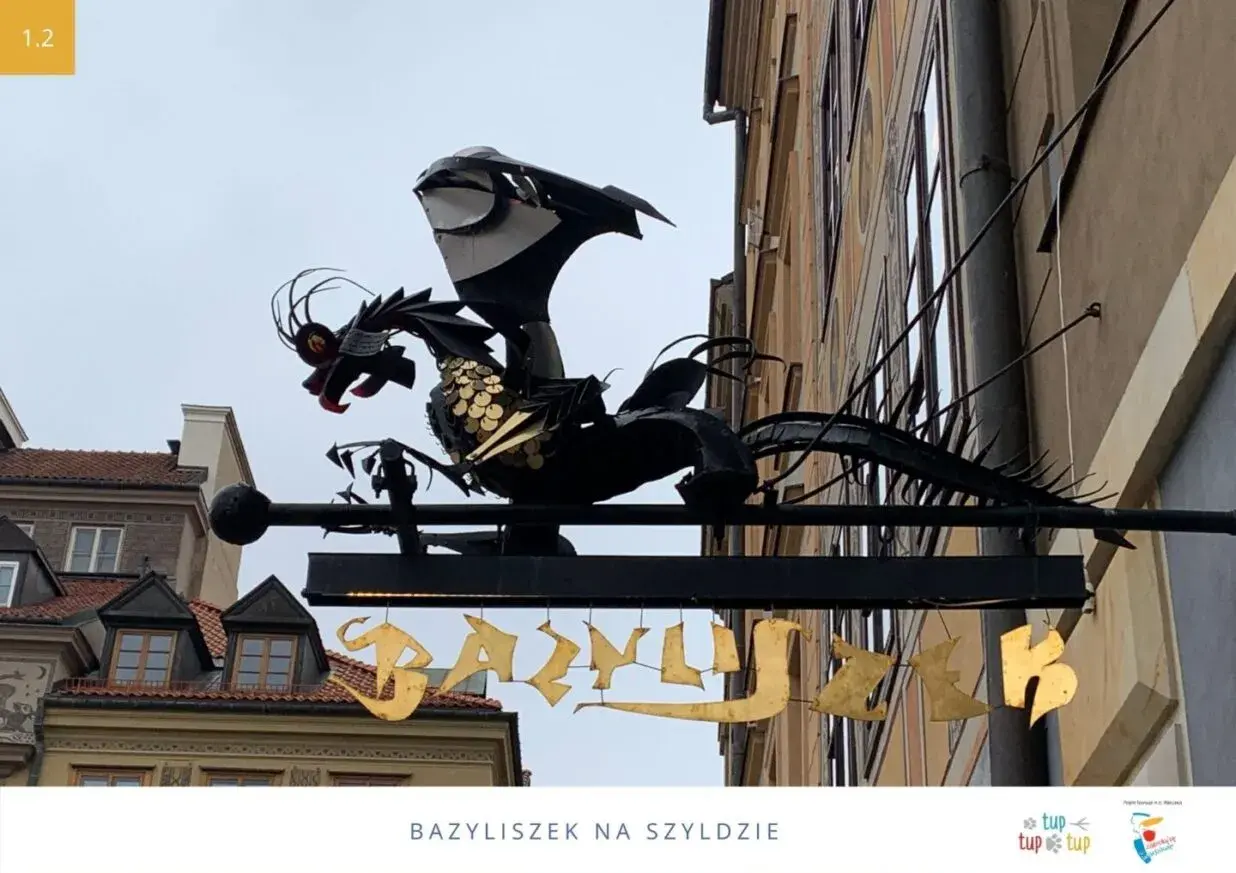 Metalowy bazyliszek ziejący ogniem wisi nad złotym napisem.