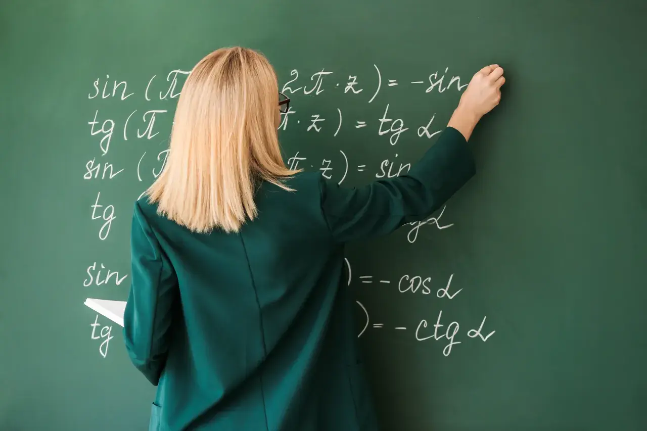 Nauczycielka pisze kredą wzory matematyczne na zielonej tablicy.