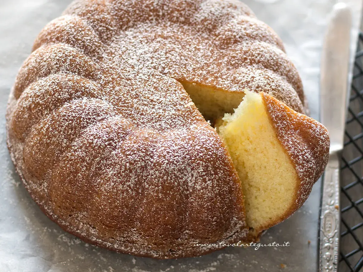 Una soffice torta allo yogurt, cosparsa di zucchero a velo, con una fetta tagliata che ne rivela l'interno dorato e profumato.