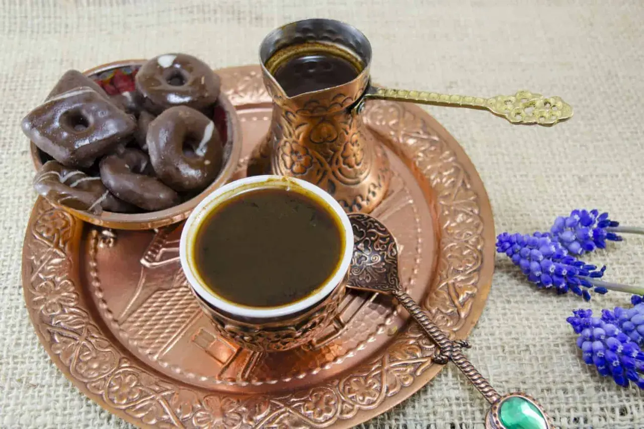 Ein traditionelles Mokkakännchen und eine Tasse mit starkem Mokka-Kaffee, dazu Schokoladenkekse und blaue Blumen auf einem Kupfertablett.