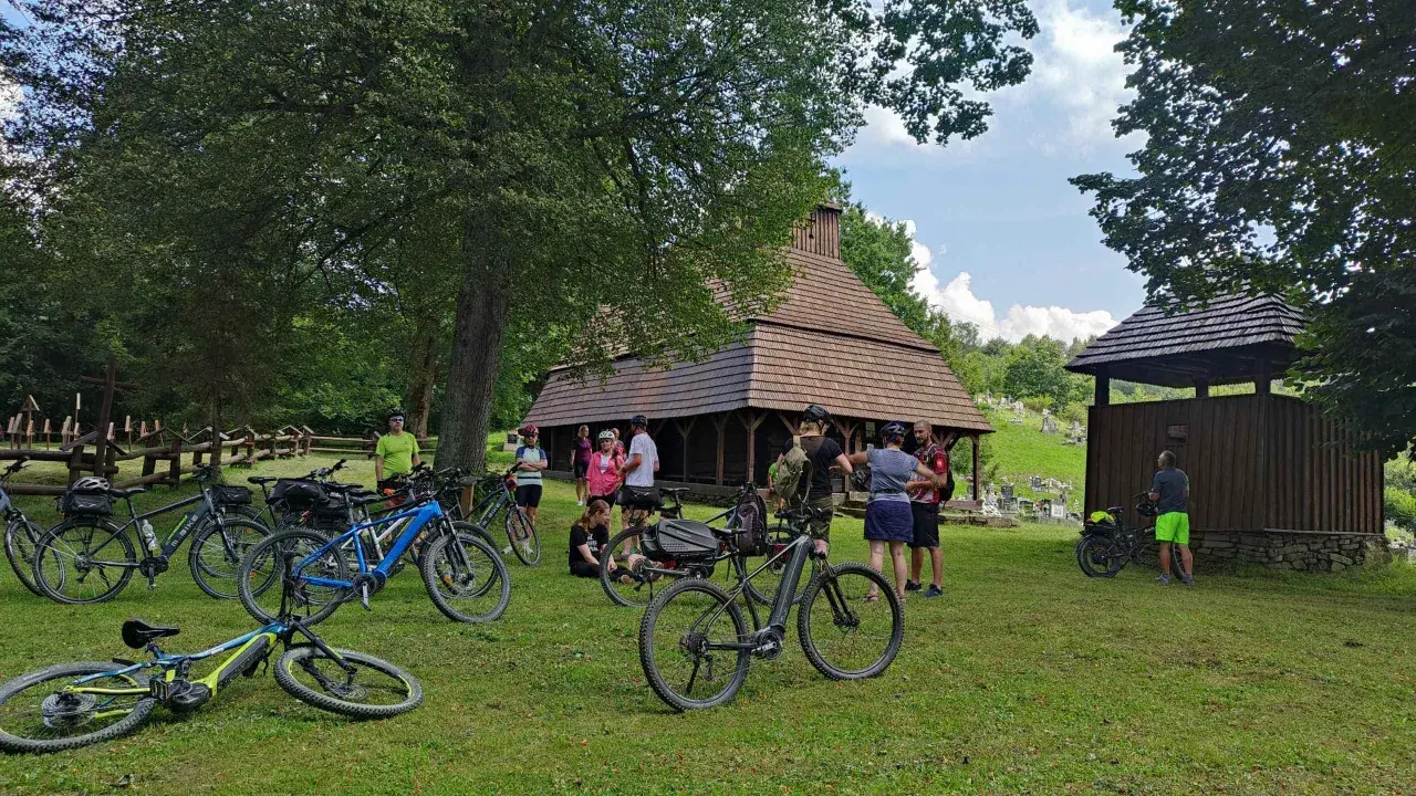 Grupa rowerzystów odpoczywa przy drewnianej cerkwi, planując dalszą podróż po malowniczych trasach rowerowych Bieszczad.