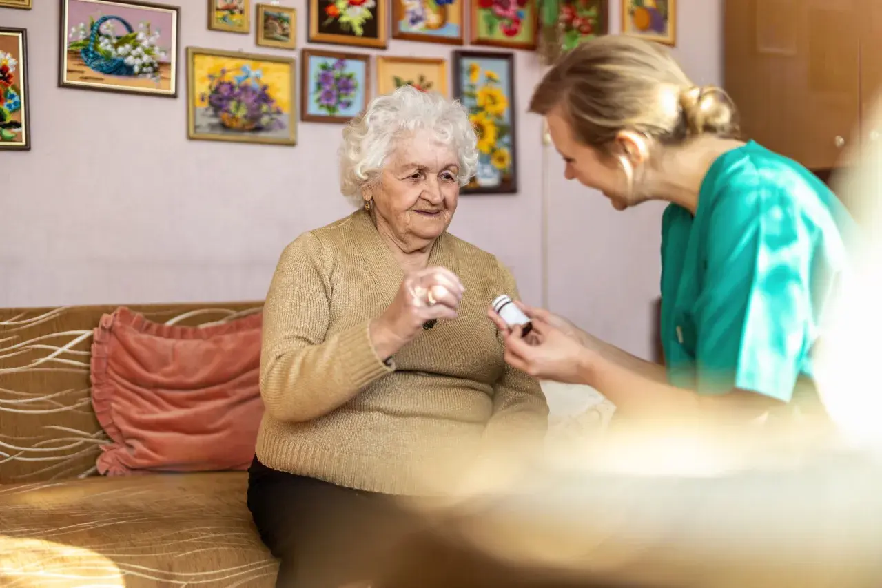 Pflegekraft reicht einer älteren Dame Medikamente für die Obstipationsprophylaxe.