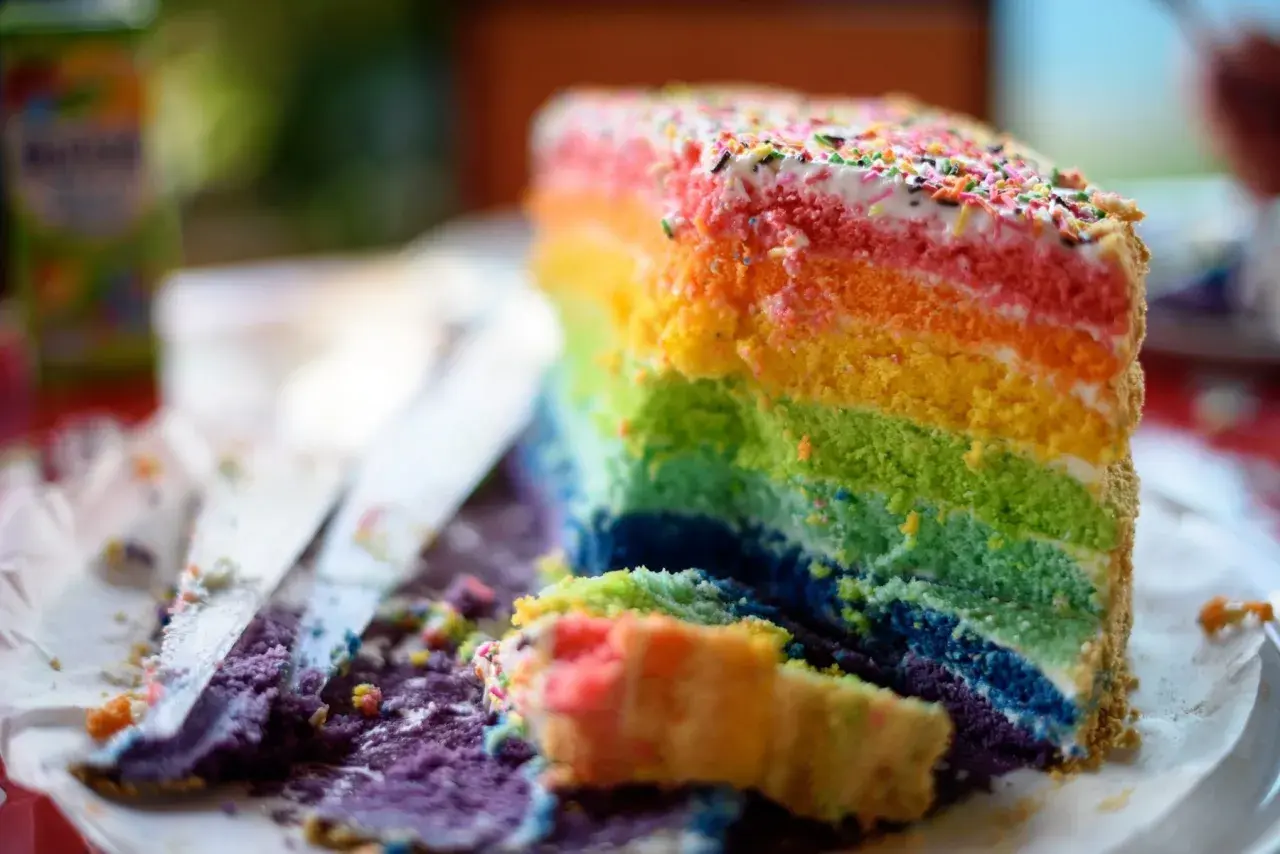 Ein Stück bunter regenbogen kuchen mit Streuseln, das auf einem Teller liegt. Daneben liegen zerbröselte Kuchenreste und ein Messer.