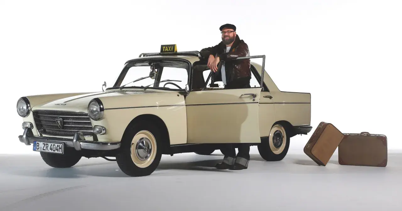 Mężczyzna w skórzanej kurtce opiera się o zabytkowy Peugeot 404 z tabliczką TAXI. Obok stoją walizki.