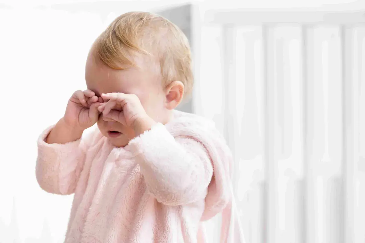 Un neonato con un pigiamino rosa strofina gli occhi, forse un occhio che lacrima per la stanchezza o un piccolo pianto.