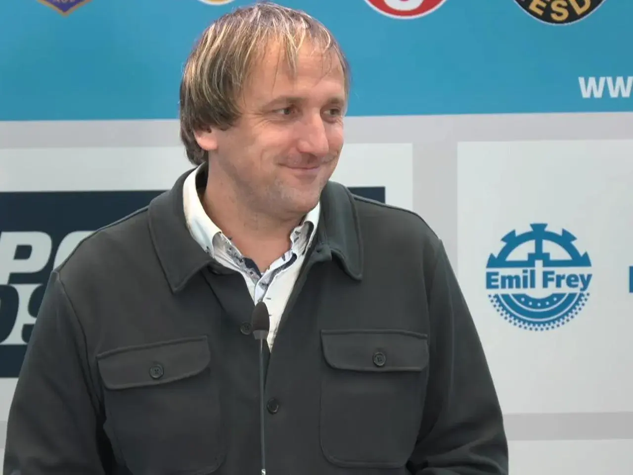 Thomas Neubert lächelt in die Kamera, trägt eine dunkle Jacke und ein gemustertes Hemd. Im Hintergrund sind Logos zu sehen.