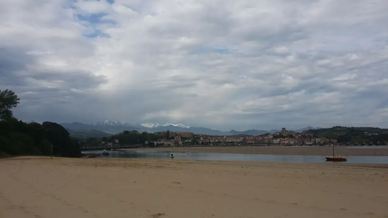 Arena dorada en la playa de la Maza, con un velero rojo en el agua y un pueblo costero al fondo, bajo un cielo nublado.