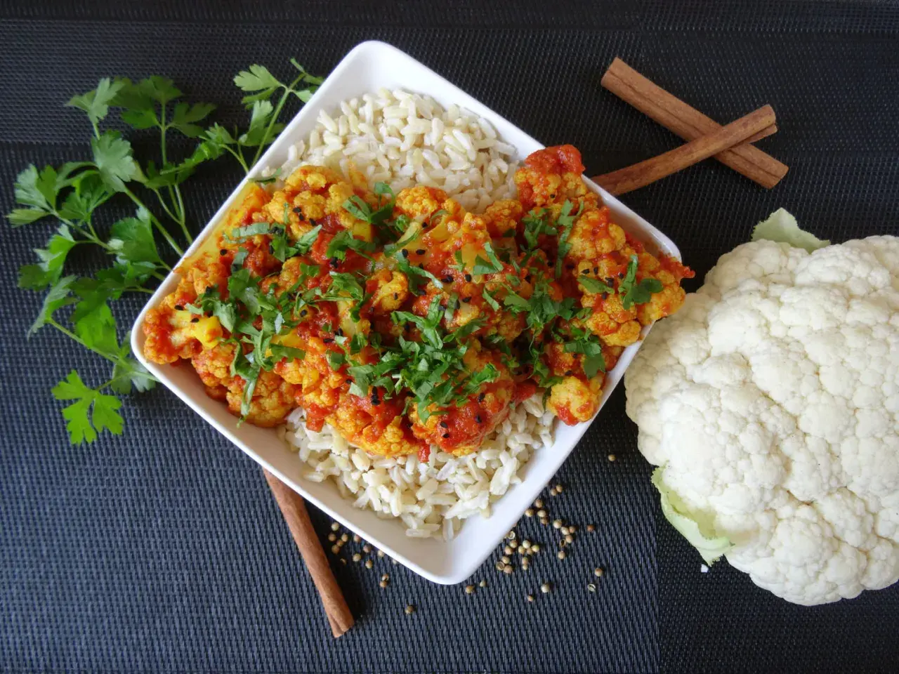 Aksamitne curry z kalafiora podane z ryżem, udekorowane świeżą natką pietruszki i czarnuszką. Obok leży świeży kalafior i laski cynamonu.