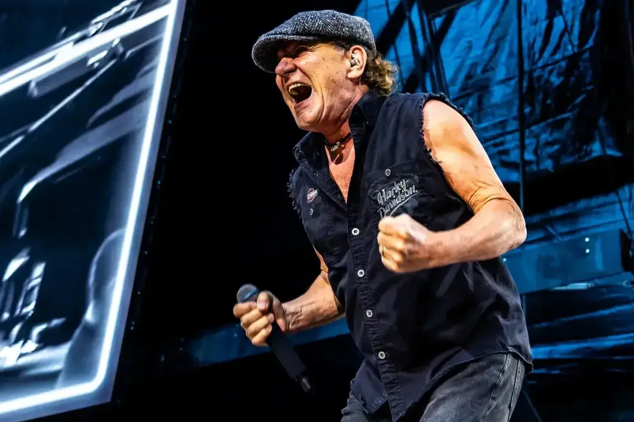 Brian Johnson z zespołu AC/DC na koncercie, w czapce i kamizelce Harley-Davidson, śpiewa z pasją do mikrofonu.