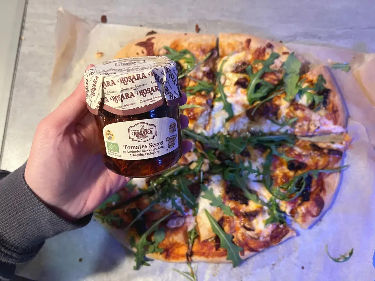Mano sostiene un tarro de tomates secos en aceite de oliva virgen extra, listo para añadir a una pizza con rúcula.