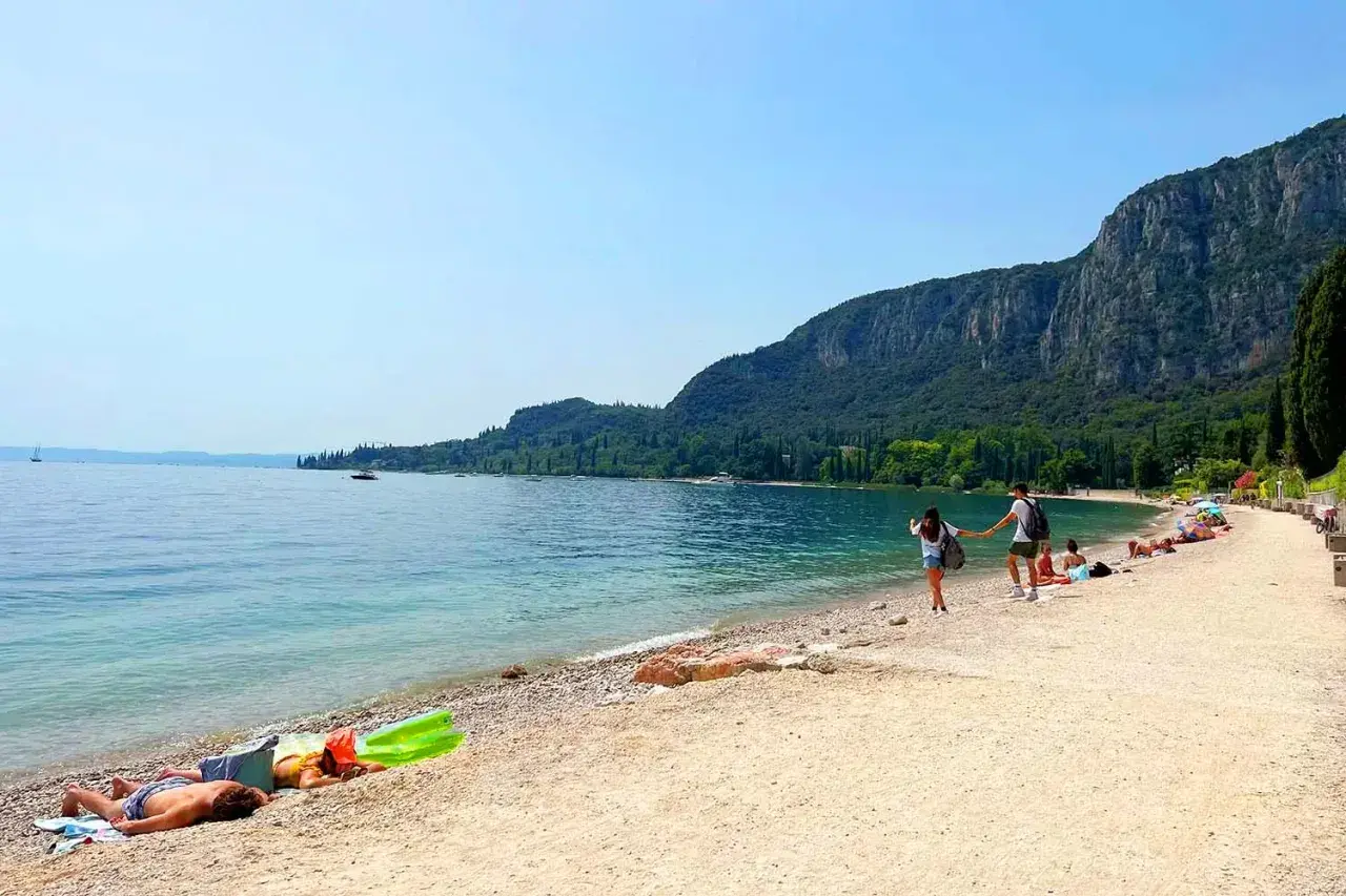 Ein Paar hält Händchen am Strand, während andere am Gardasee schwimmen. Berge und Bäume säumen die Küste.