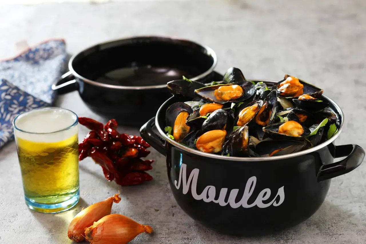 Un bicchiere di birra dorata accanto a cozze fumanti in una pentola nera, con peperoncini secchi e scalogni. Un perfetto abbinamento di birra e pesce.