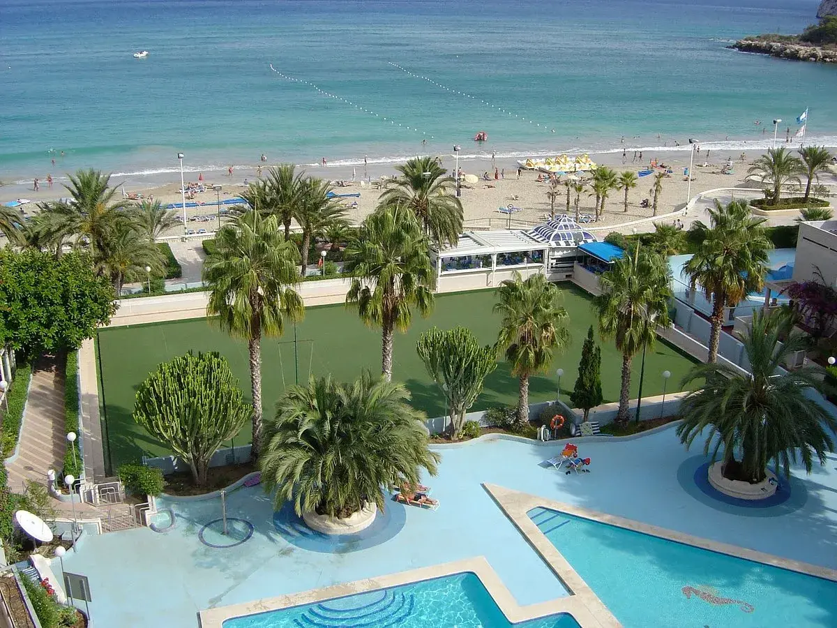 Vista aérea de un resort con piscina, cancha de césped, palmeras y una playa con el mar azul. Un lugar perfecto para sol y mar spa.