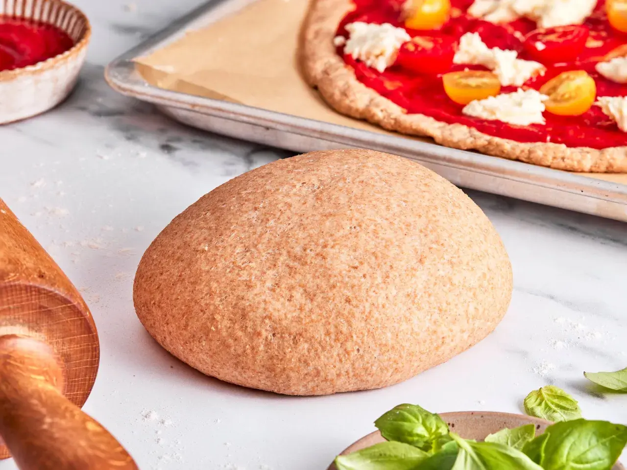 Ein runder Laib vollkorn pizzateig liegt bereit. Im Hintergrund eine fast fertige Pizza mit Tomaten und Mozzarella.