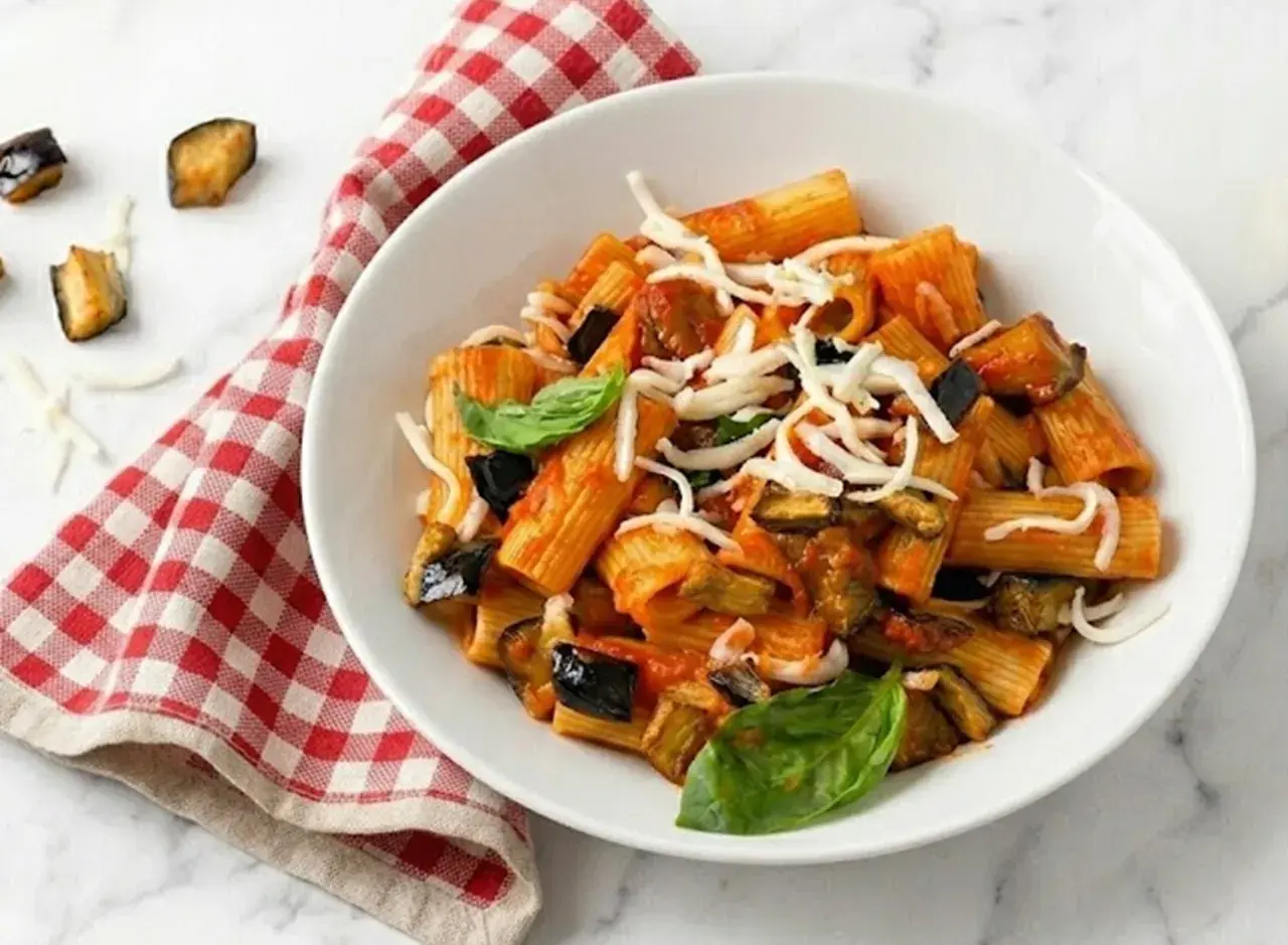 Un plato de pasta alla norma con berenjenas fritas, salsa de tomate y queso rallado, decorado con albahaca fresca.