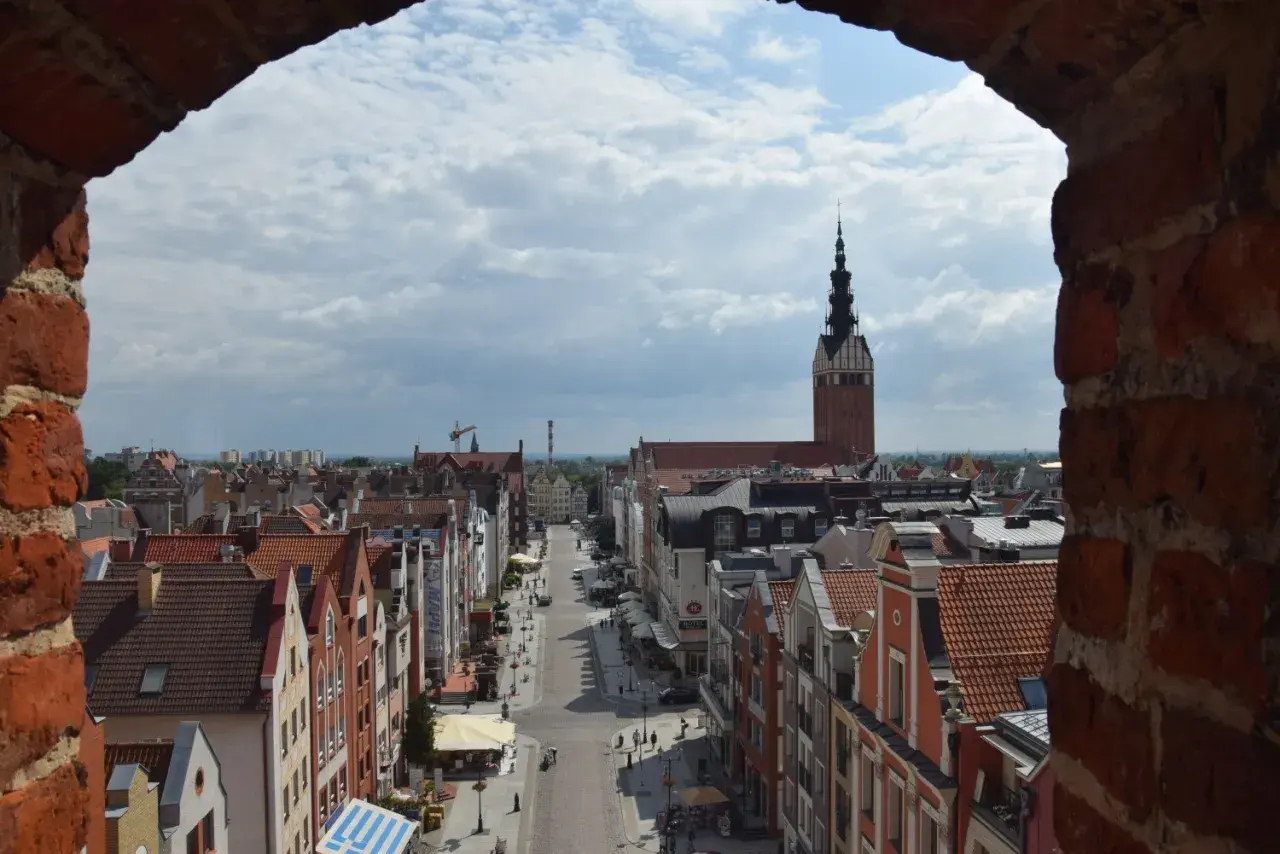 Widok na ulicę i kamienice w **starym mieście Elbląg**. W oddali widać wieżę kościoła.