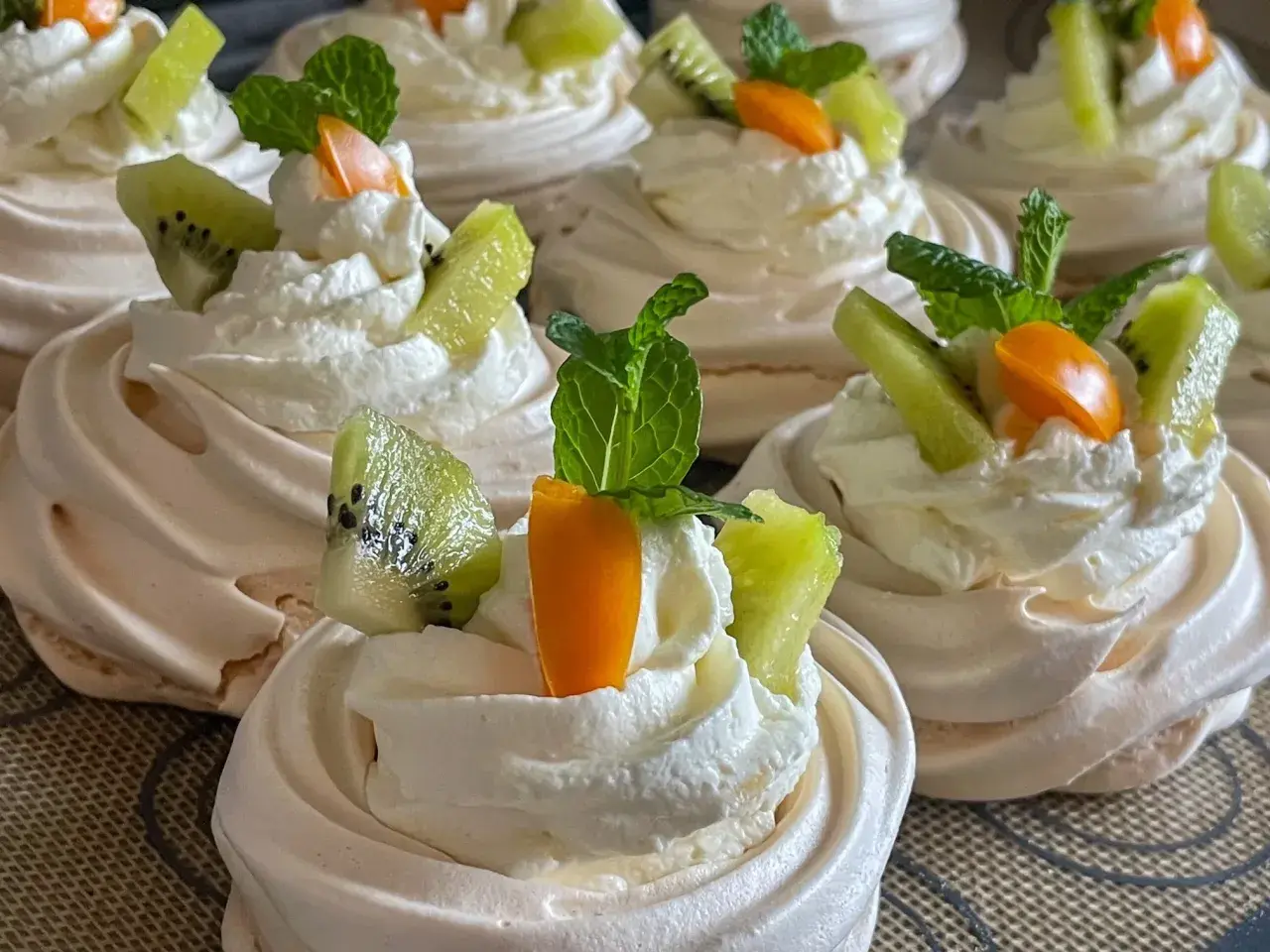 Pyszne gniazdka bezowe z kremem mascarpone, ozdobione kawałkami kiwi, pomarańczy i listkami mięty.