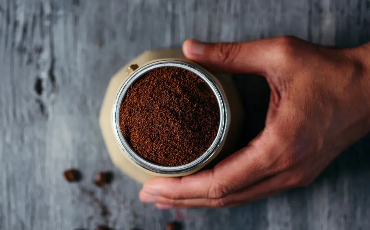 Mano che tiene la moka piena di caffè macinato, pronto per il rito del caffè. Chicchi sparsi sul tavolo di legno.
