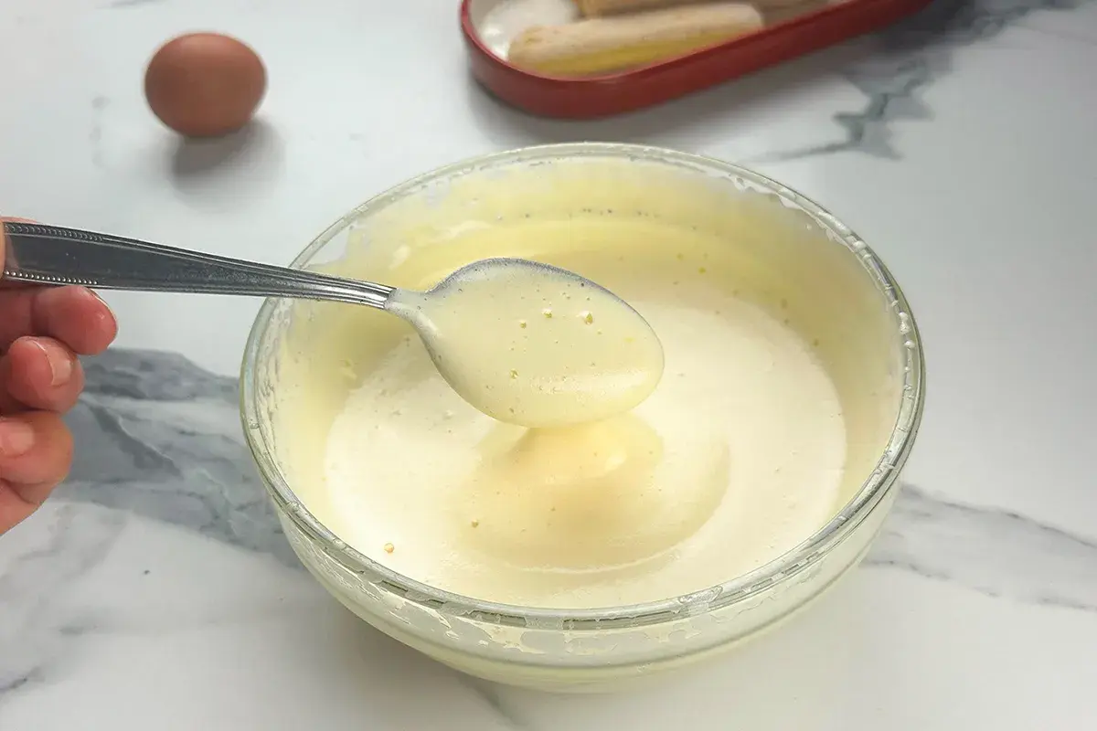 Préparation d'un tiramisu : une cuillère de pâte à bombe aérienne est prélevée d'un bol en verre. Un œuf et des biscuits à la cuillère sont visibles.