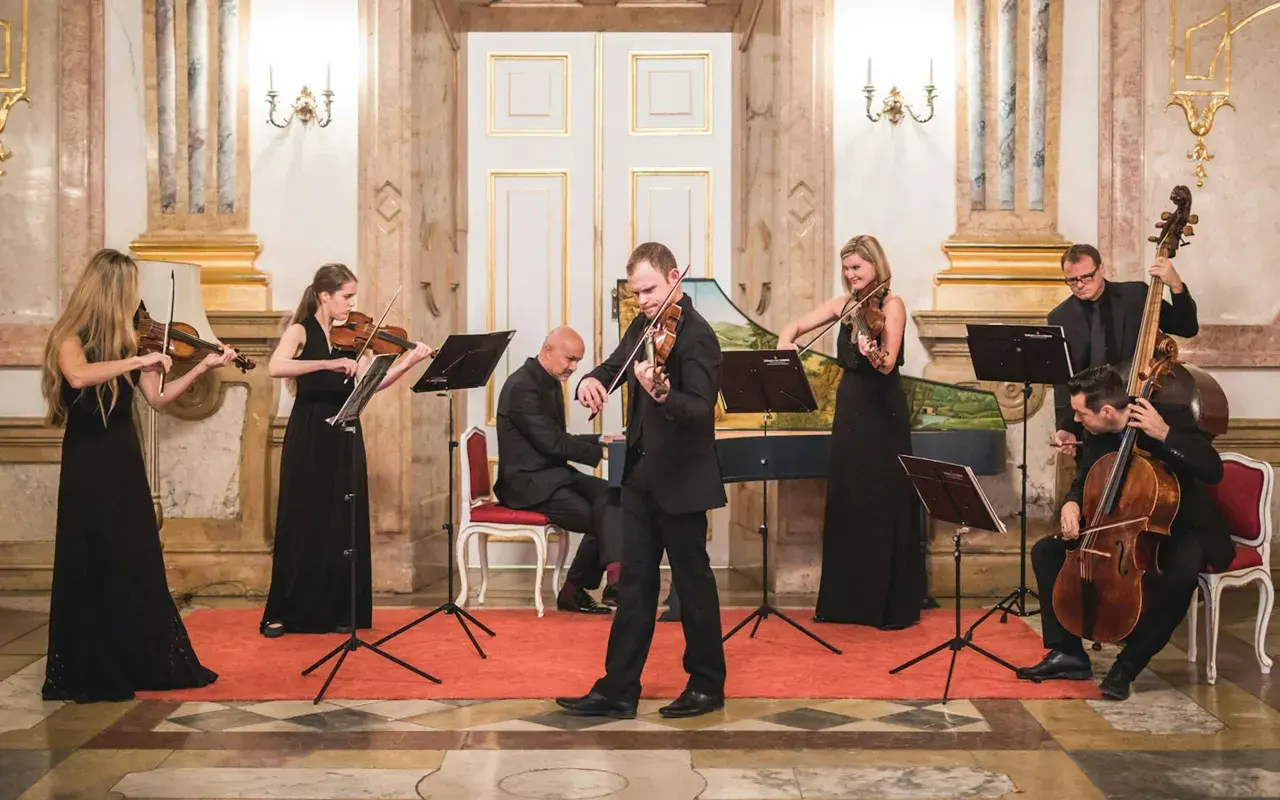 Ensemblen ger en klassisk musik konsert i en elegant sal. Sex musiker spelar fioler, cello och cembalo.