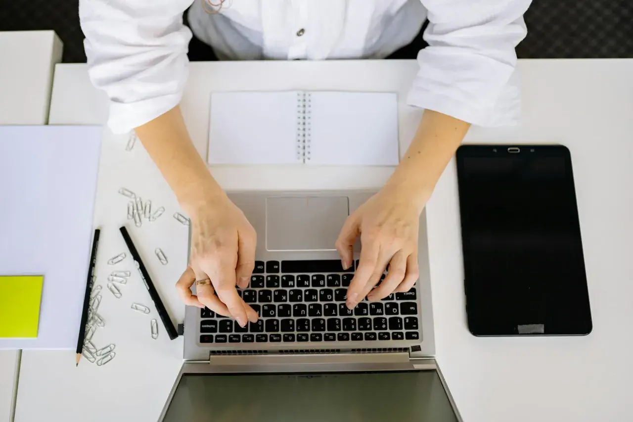 Kobieta pisze na laptopie, szukając informacji, jak napisać stopnie Celsjusza w Excelu. Obok leżą tablet, notatnik i spinacze.
