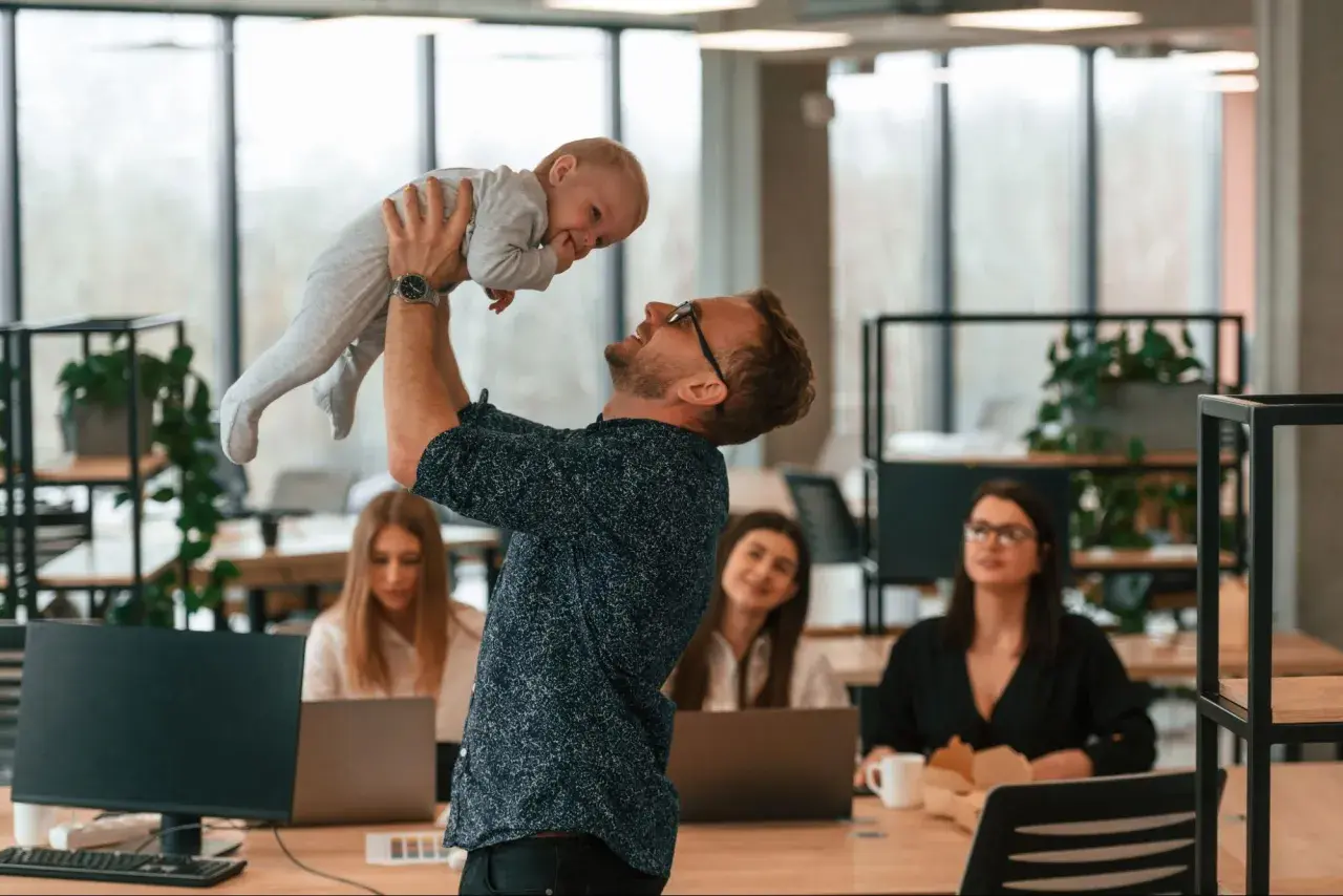 Mężczyzna w okularach podnosi dziecko do góry w biurze, w tle pracują kobiety.