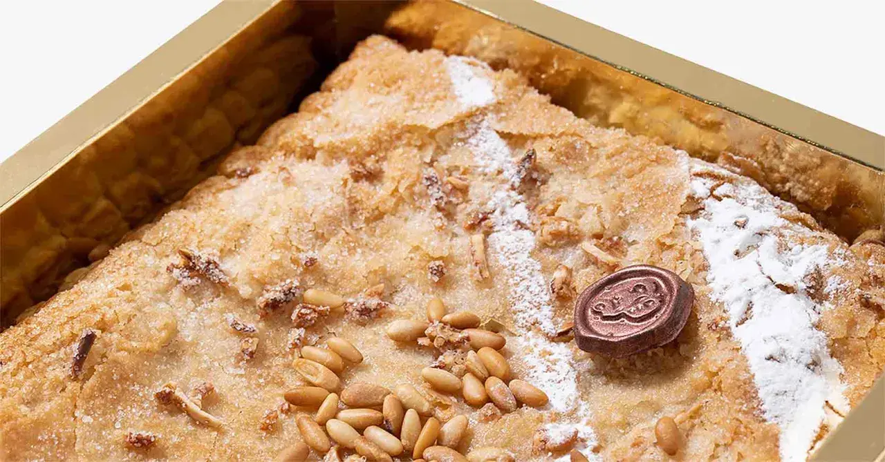 Deliciosa coca de Llavaneres con piñones y un toque de azúcar glas, presentada en una caja dorada.