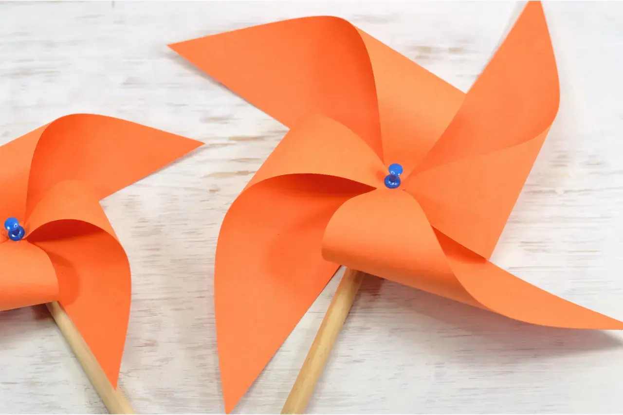 Zwei selbstgebastelte Windräder aus orangefarbenem Papier auf Holzuntergrund. Perfekt zum Windrad basteln mit Kindern.