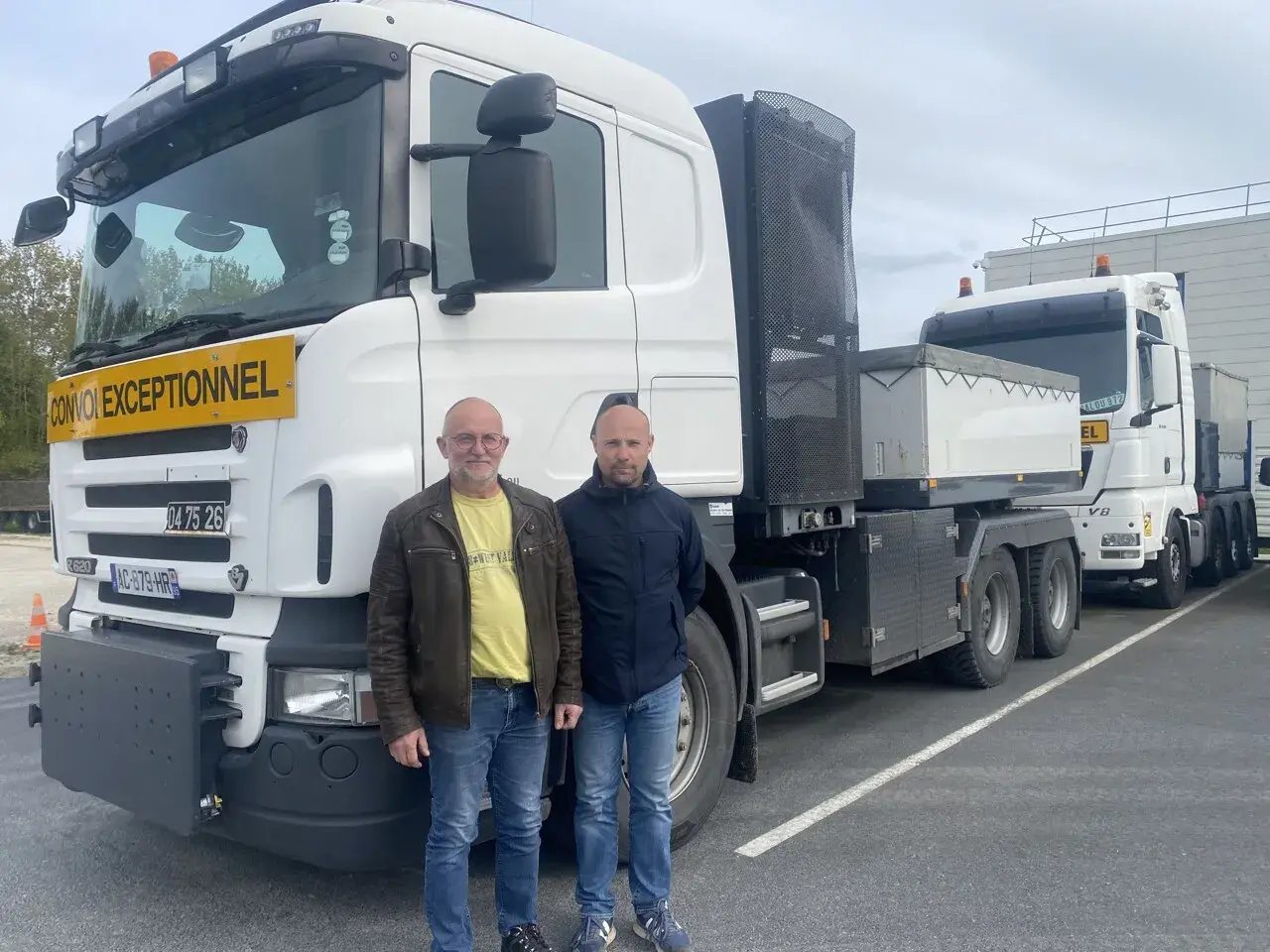 Deux chauffeurs poids lourd national posent devant leurs camions. L'un porte un gilet jaune, l'autre une veste bleue.