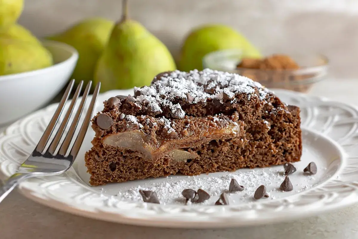 Una fetta di torta pere e cioccolato, cosparsa di zucchero a velo e gocce di cioccolato, con pere caramellate all'interno.